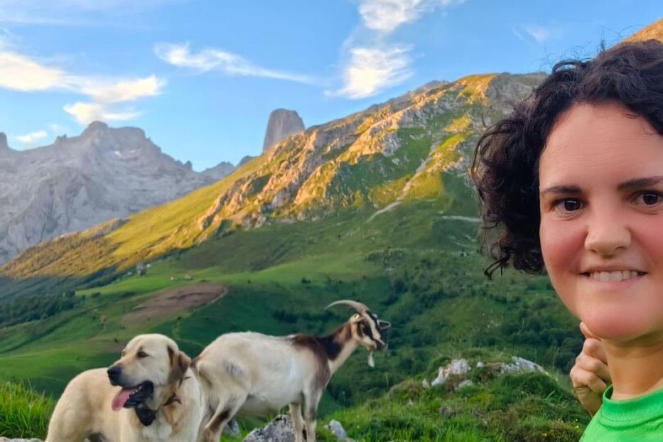 Eine Frau macht ein Selfie mit einem Hund und einer Ziege vor einer sonnigen Bergkulisse.