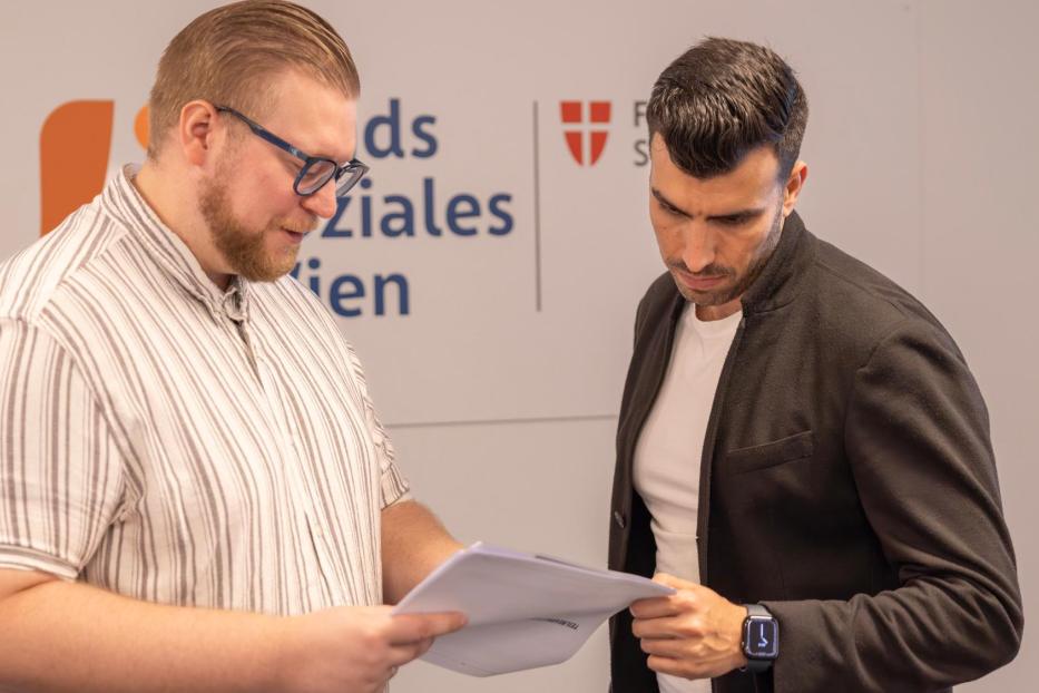 Zwei Männer besprechen Unterlagen vor dem Fonds-Soziales-Wien-Logo.