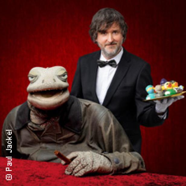 Ein Mann im Anzug präsentiert eine Echsenpuppe mit Zigarre und ein Tablett mit Süßigkeiten.