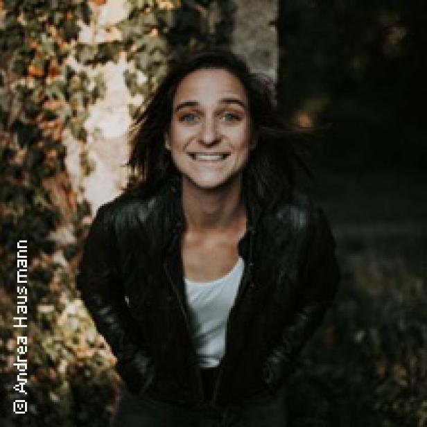 Eine lächelnde Frau in Lederjacke steht vor einer mit Efeu bewachsenen Wand.