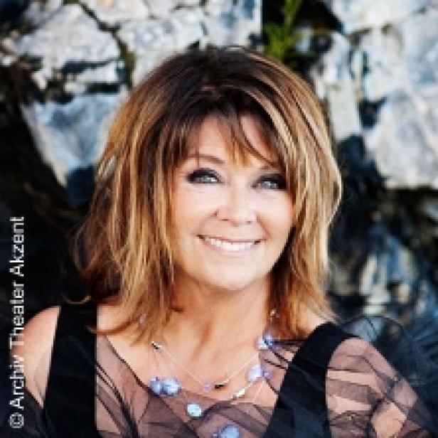 Eine lächelnde Frau mit braunen Haaren und einer Halskette vor einem Felsen.