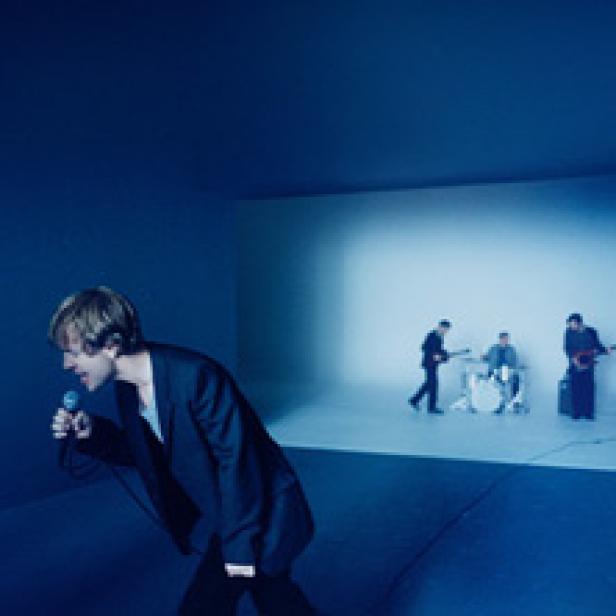 Eine Band spielt in einem blauen Raum, wobei der Sänger im Vordergrund steht.