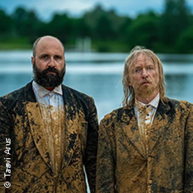 Zwei Männer in schlammverschmierten Anzügen stehen vor einem See.
