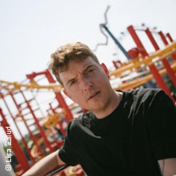 Ein Mann mit blonden Haaren vor einer Achterbahn.
