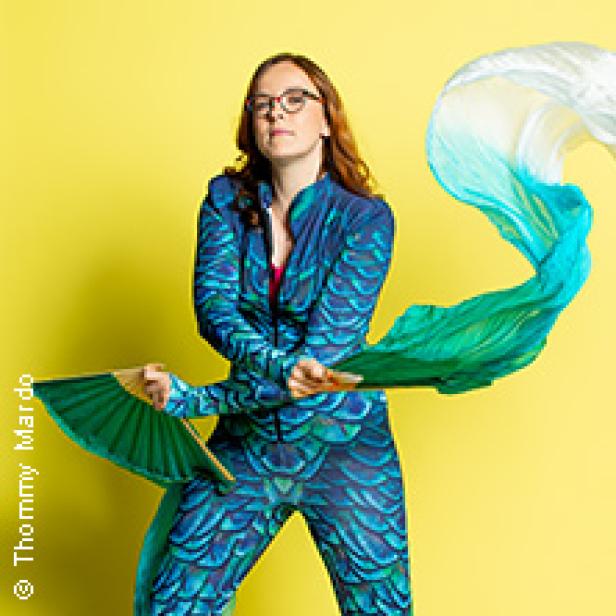 Eine Frau in einem blaugrünen Kostüm posiert mit einem Fächer und einem wehenden Tuch vor einem gelben Hintergrund.