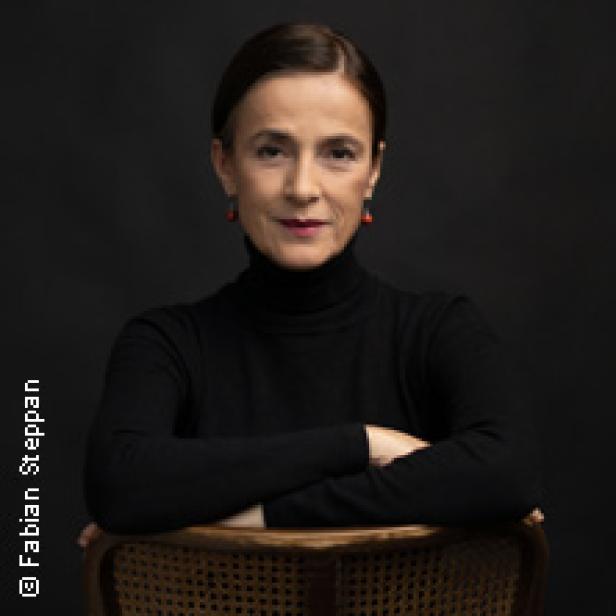 Eine Frau mit dunklen Haaren und einem schwarzen Rollkragenpullover sitzt auf einem Stuhl.