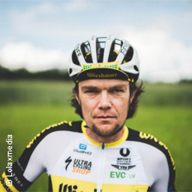 Ein Radfahrer in Sportkleidung und Helm steht vor einer grünen Landschaft.