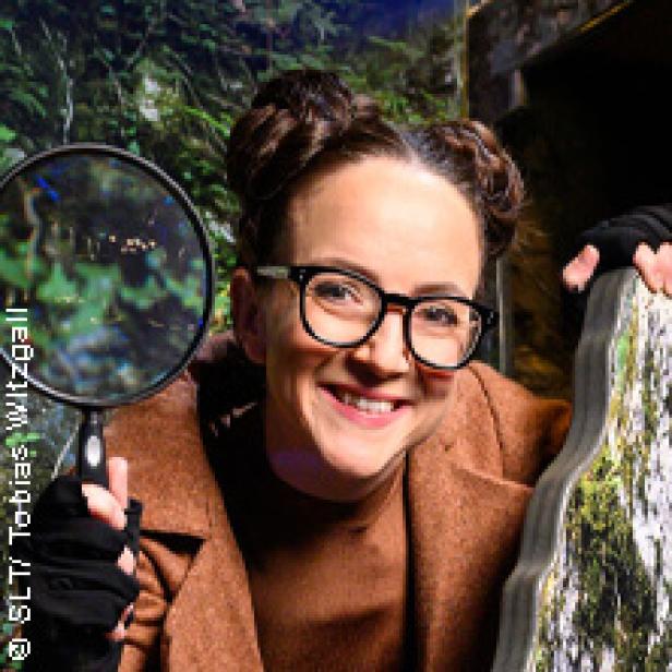 Eine lächelnde Frau mit Brille und Dutt hält eine Lupe vor einem grünen Hintergrund.