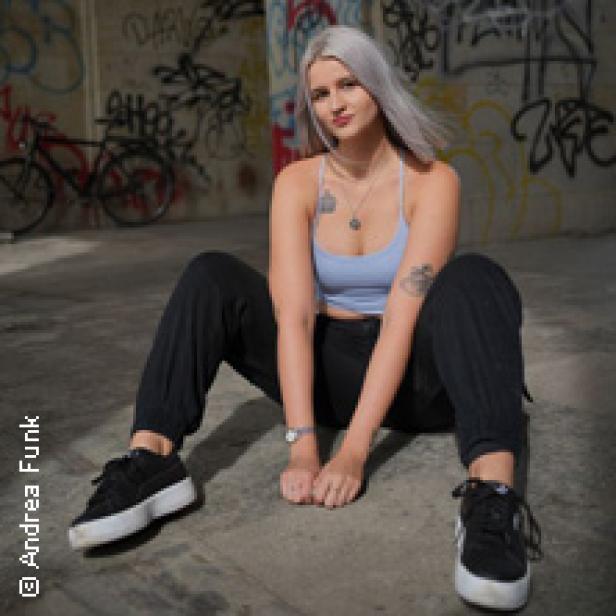 Eine junge Frau mit hellblondem Haar sitzt vor einer Graffitiwand.
