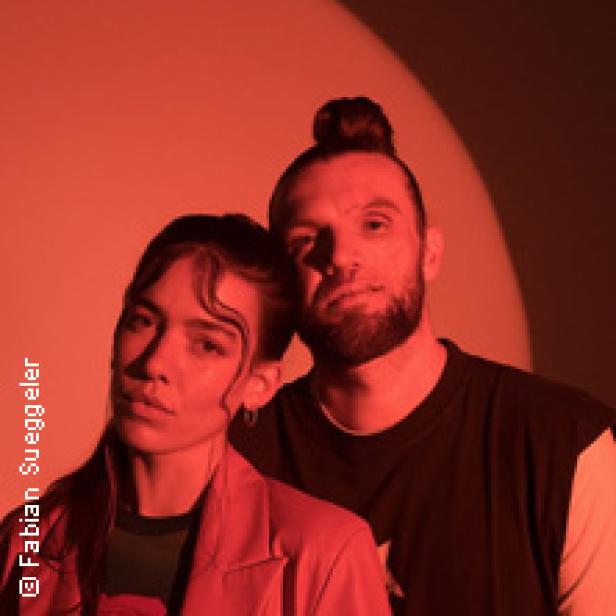 Ein Mann mit Dutt und eine Frau mit Blazer posieren vor einem roten Hintergrund.