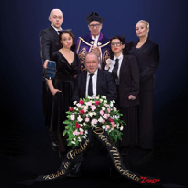 Eine Gruppe von Personen in Trauerkleidung steht um einen Kranz mit der Aufschrift „Ruhe in Frieden, Deine Mutter Mitarbeiter Team“.