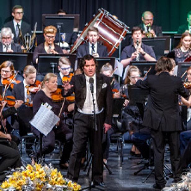 Ein Sänger steht vor einem Orchester und singt in ein Mikrofon.