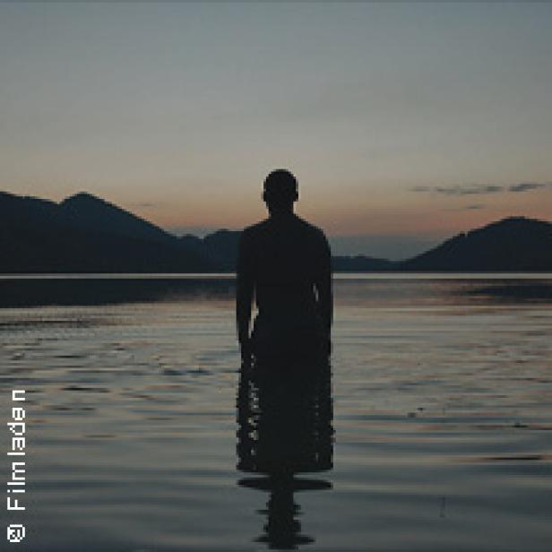 Eine Person steht im Wasser vor einer Bergkulisse im Abendlicht.