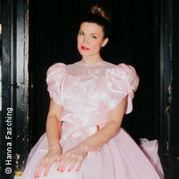 Eine Frau in einem rosa Kleid sitzt vor einem dunklen Hintergrund.