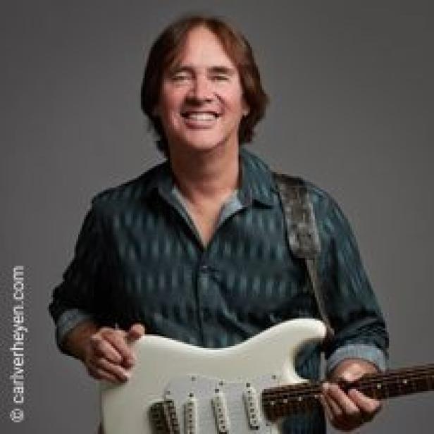 Carl Verheyen hält eine weiße E-Gitarre und lächelt.
