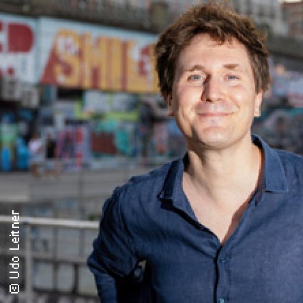 Ein Mann mit blauem Hemd lächelt vor einer Wand mit Graffiti.