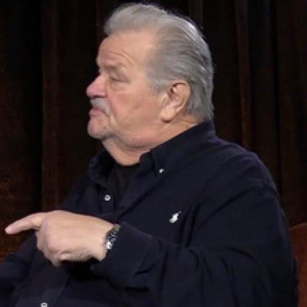 Ein Mann mit grauem Haar und Schnurrbart zeigt mit dem Finger nach links.