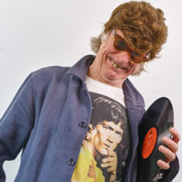 Ein Mann mit Perücke und Sonnenbrille lächelt und hält eine Schallplatte.