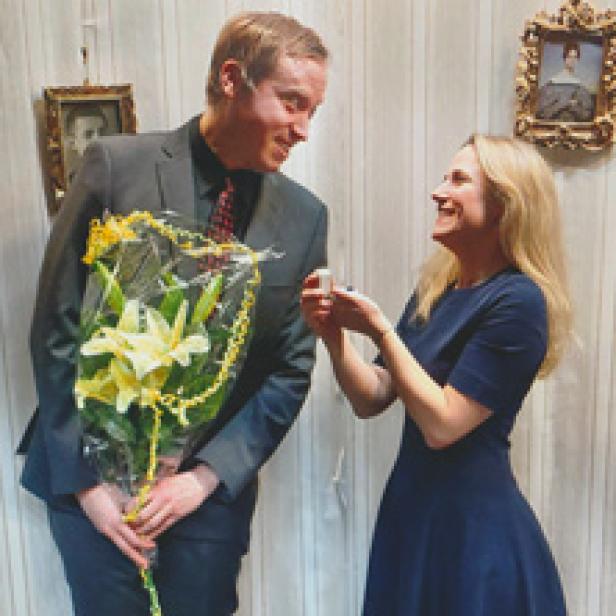 Ein Mann mit Blumenstrauß hält um die Hand einer lachenden Frau an.