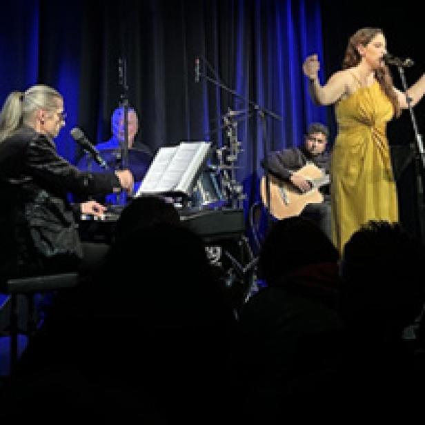 Eine Sängerin tritt mit ihrer Band auf einer Bühne vor einem dunklen Vorhang auf.