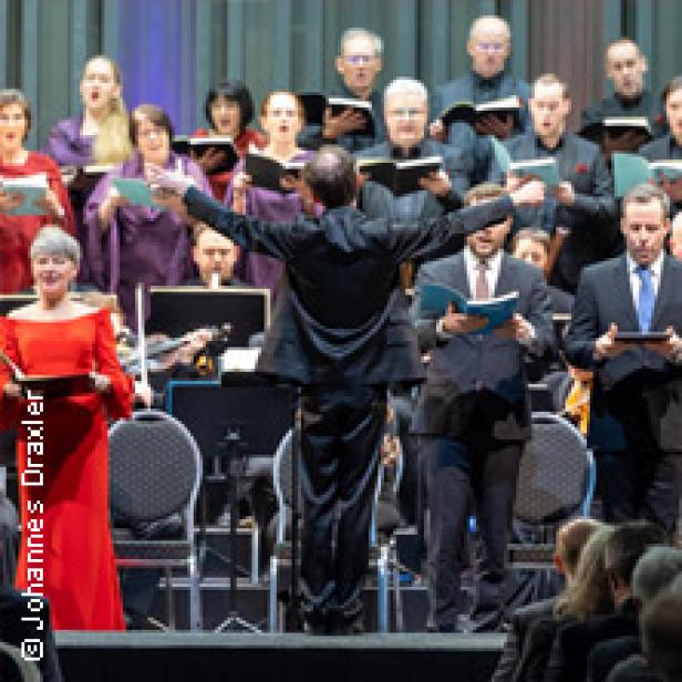Ein Dirigent steht vor einem Chor und einem Orchester während einer Aufführung.