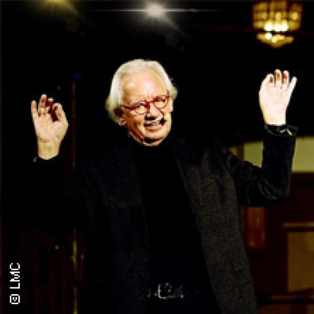 Ein älterer Mann mit Brille gestikuliert mit erhobenen Händen.