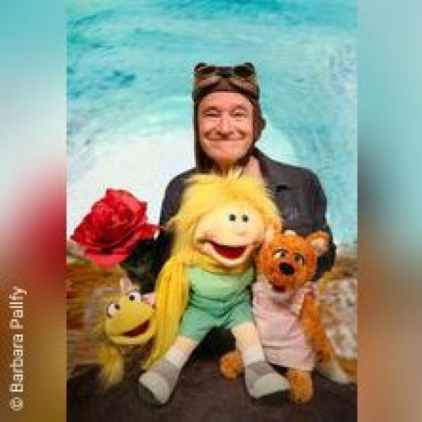 Ein Mann mit Fliegerbrille posiert mit vier bunten Handpuppen und einer roten Rose vor einem hellblauen Hintergrund.