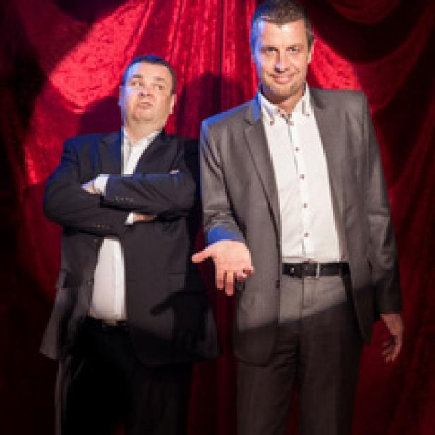 Zwei Männer in Anzügen stehen vor einem roten Vorhang.