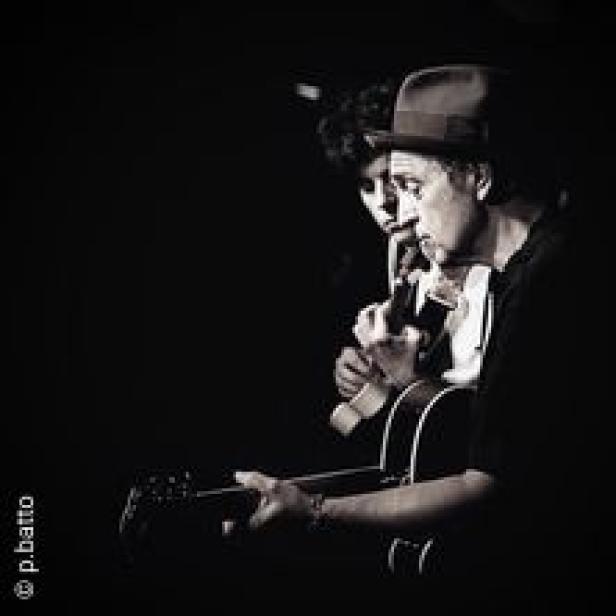 Ein Mann mit Hut spielt Gitarre, während eine Frau ihm aufmerksam zuhört.