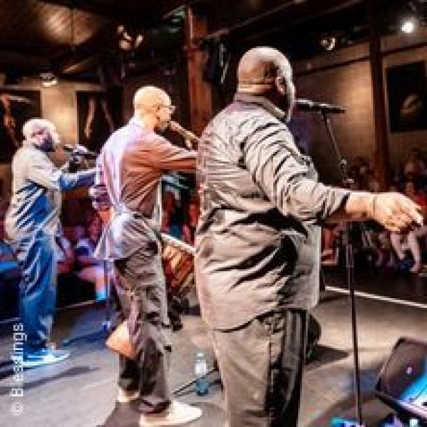 Drei Musiker stehen auf einer Bühne und spielen vor einem Publikum.