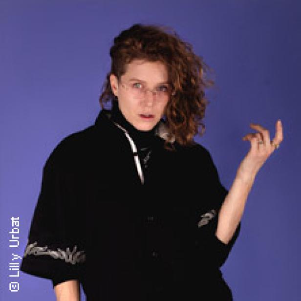 Eine Frau mit lockigem Haar und schwarzer Jacke posiert vor einem blauen Hintergrund.