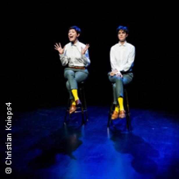 Zwei Personen sitzen auf Stühlen und scheinen auf einer Bühne zu performen.