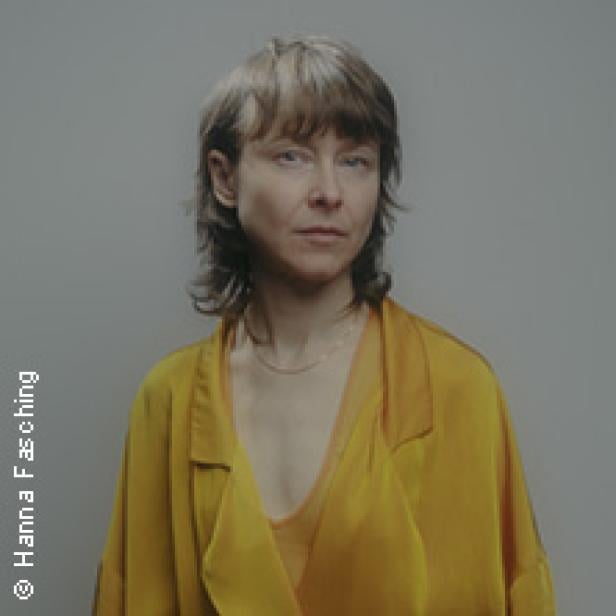 Porträt einer Frau mit blonden Haaren und gelber Bluse vor einem grauen Hintergrund.