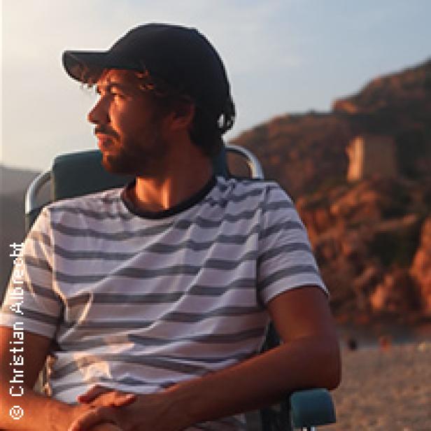 Ein Mann mit Bart und Kappe sitzt am Strand und schaut in die Ferne.