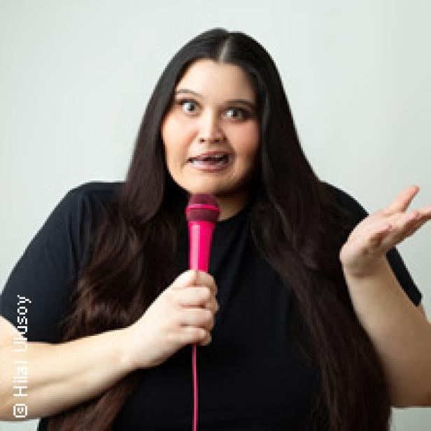 Eine Frau mit langen, dunklen Haaren hält ein pinkfarbenes Mikrofon und spricht.