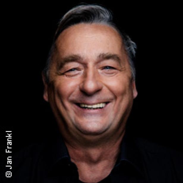 Ein lächelnder Mann mit kurzen, grauen Haaren vor einem schwarzen Hintergrund.