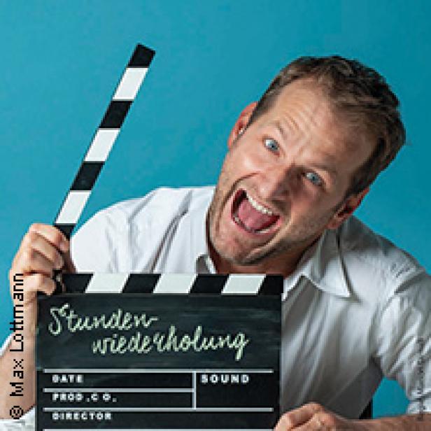 Ein Mann hält eine Filmklappe mit dem Titel „Stundenwiederholung“ in die Kamera.