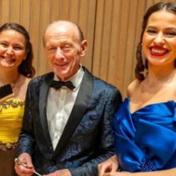 Ein älterer Mann in einem Anzug posiert mit zwei jungen Frauen in Abendkleidern.