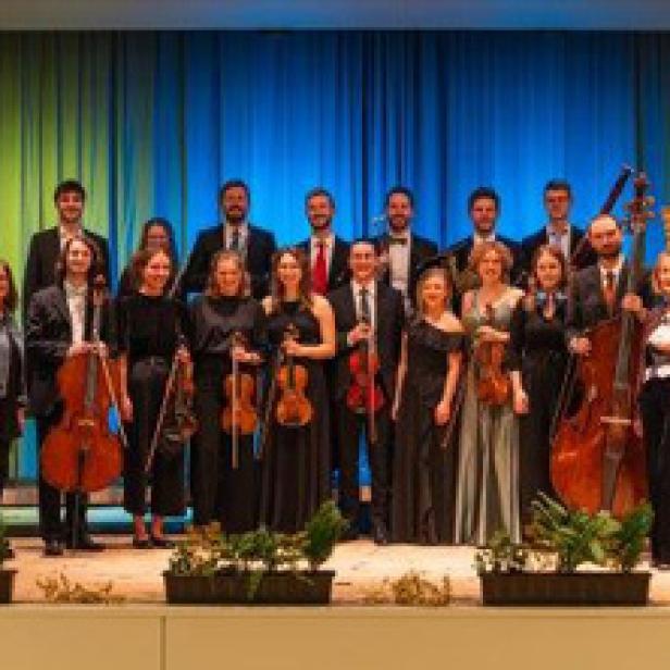 Ein Orchester steht auf einer Bühne vor einem blauen Vorhang.