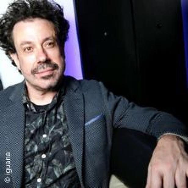 Ein Mann mit dunklen, lockigen Haaren und Bart trägt ein dunkles Sakko.