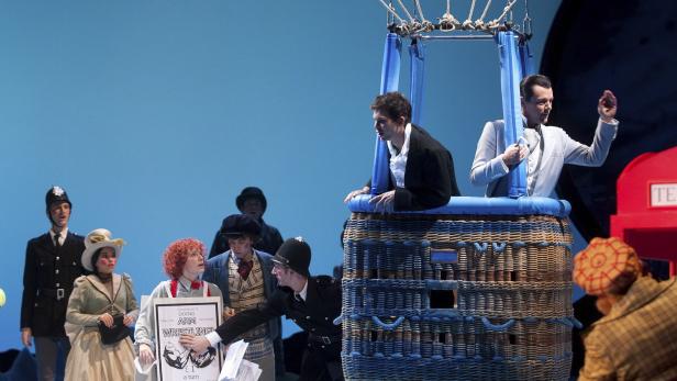 Eine Theaterszene mit Menschen in Kostümen, einem Heißluftballon und einer Telefonzelle.