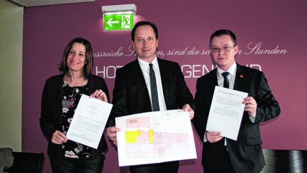 Drei Personen präsentieren Dokumente und einen Plan vor einer Wand mit Schriftzug.