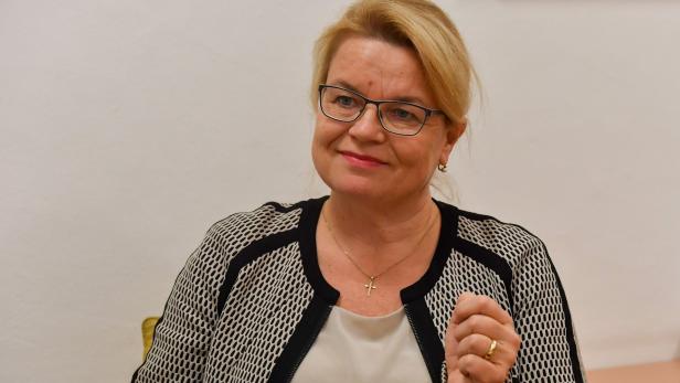 Eine blonde Frau mit Brille und Kreuzkette lächelt.