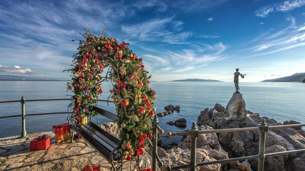 Ein mit Weihnachtsschmuck dekorierter Bogen und Geschenke vor einer Küstenlandschaft mit einer Statue.