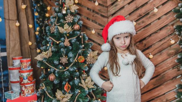 Disappointed little girl wearing Christmas costume standing isolated over wooden christmas background, keeping arms akimbo, frowning her face.