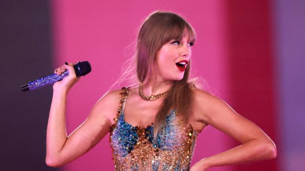 Taylor Swift auf der Bühne mit einem Mikro in der Hand, schaut verschmitzt