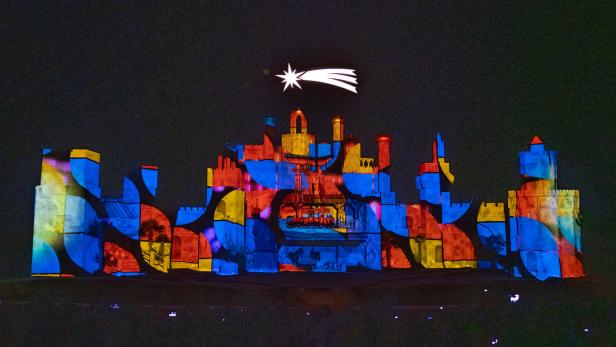 Eine farbenfrohe Projektion auf eine Burgmauer unter einem hellen Sternenhimmel.