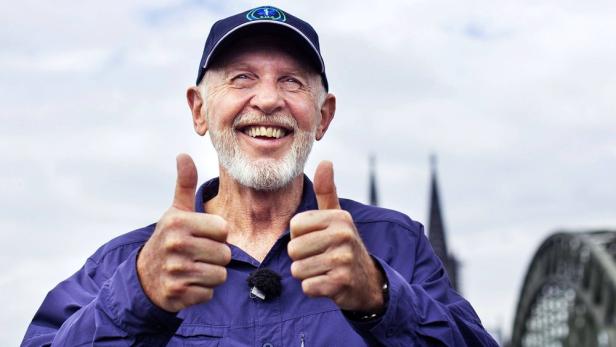 Dr. Bob vom Dschungelcamp lächelt und zeigt beide Daumen nach oben. Im Hintergrund sind Domspitzen zu sehen.