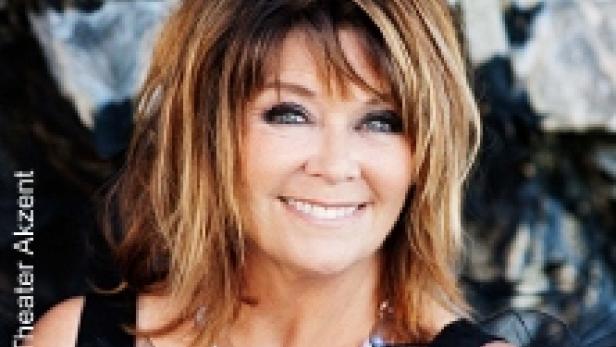 Eine lächelnde Frau mit braunen Haaren und einer Halskette vor einem Felsen.