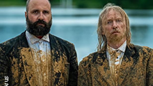 Zwei Männer in schlammverschmierten Anzügen stehen vor einem See.